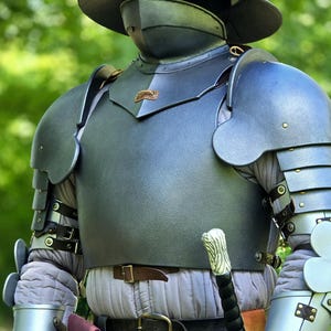 Könnte beinhalten: Eine Rüstung aus Stahlplatten mit einem Kesselhut-Helm mit Gesichtsschutz, einem Brustpanzer, beweglichen Schulterstücken und Panzerhandschuhen. Die Rüstung wird über einem hellgrauen gesteppten Gambeson getragen und umfasst ein Schwert mit weißem Griff.