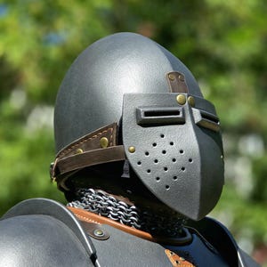 May include: A matte black helmet with a pointed visor featuring horizontal eye slits and a perforated ventilation section. The helmet is secured with brown leather straps and brass rivets, worn over a metallic chainmail neck guard and armor.