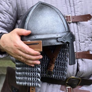 May include: A medieval-style helmet with a chainmail neck guard is held against a gray quilted jacket. The helmet is dark gray with a curved top and a visor. Brown leather straps and a brown leather pouch are also visible.