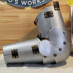 Könnte beinhalten: Silberfarbene Armrüstung mit genieteten braunen Lederakzenten. Die Rüstung ist am Ellbogen und Handgelenk beweglich. Der Hintergrund zeigt ein Schild mit der Aufschrift "Niko's Workshop" und eine Holzoberfläche.