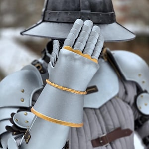 Può includere: Un'armatura medievale, principalmente grigio argento, con dettagli dorati. L'armatura comprende un elmo, spallacci e guanti. I guanti hanno finiture dorate e una fascia dorata intrecciata. L'armatura è indossata sopra un indumento imbottito.