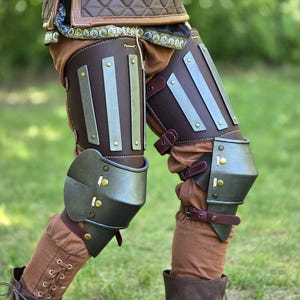 May include: A set of leg armor featuring dark brown leather thigh guards with vertical metal plates, dark grey metal knee caps, and brown fabric trousers. The armor is secured with dark brown leather straps and brass-colored rivets.