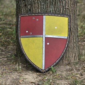 May include: A heater-style shield featuring a quartered design with alternating red and yellow panels. A central silver-coloured metal cross divides the quadrants, secured by visible rivets. The shield is finished with a dark brown leather border.