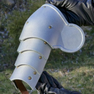 May include: Silver-colored arm armor with riveted details and a brown leather strap. The armor is segmented and appears to be made of metal. The armor is held by a person wearing black gloves.