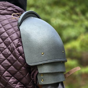 Op de afbeelding: Een grijze schouderharnas met een getextureerde afwerking, bestaande uit drie overlappende platen die zijn vastgezet met kleine messingkleurige klinknagels. Een bruine leren riem steekt uit de onderste plaat, gedragen over een paars doorgestikt kledingstuk.