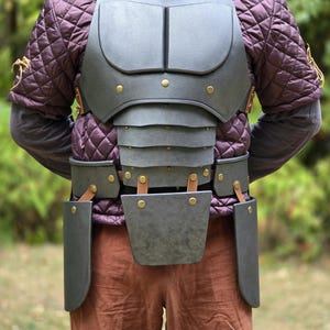 May include: A dark gray chest plate armor set featuring a segmented torso design with brass-colored rivets and leather straps. The armor is worn over a purple quilted long-sleeve garment and brown trousers, providing torso and hip protection.