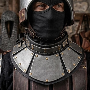 May include: A metallic helmet with a rounded crown and a black fabric face mask covering the nose and mouth. A dark leather gorget with steel plates and brass rivets protects the neck, layered over a brown leather chest piece with vertical panels.