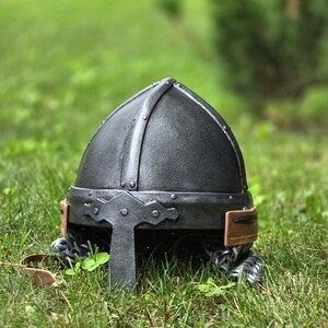 May include: A black medieval helmet with a rounded top, riveted construction, and a decorative brow plate. The helmet features a brown leather strap and chainmail around the neck. It is set against a backdrop of green grass.