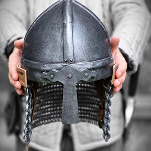 May include: A silver-colored medieval helmet with a pointed top and a face guard. The helmet features rivets, chainmail, and leather straps. The helmet is held up by a person.