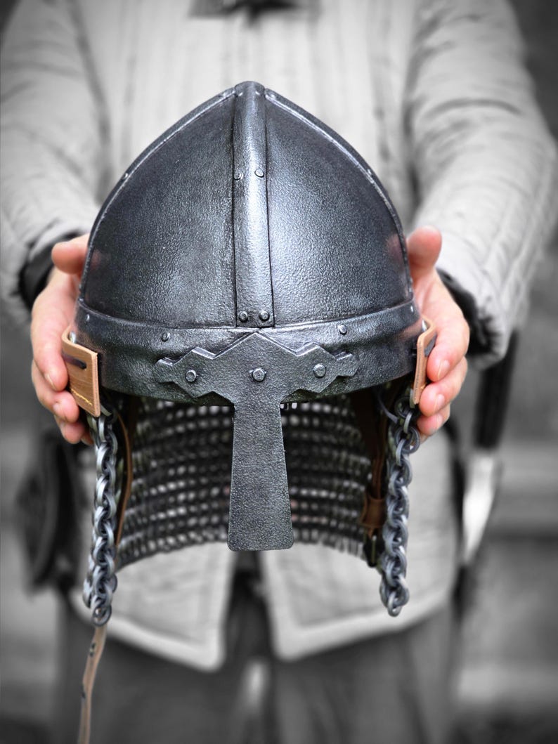 May include: A silver-colored medieval helmet with a pointed top and a face guard. The helmet features rivets, chainmail, and leather straps. The helmet is held up by a person.