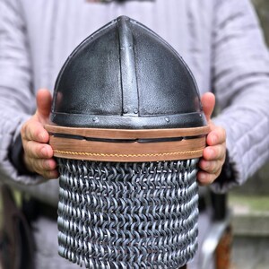 May include: A medieval-style helmet with a dark, metallic finish. The helmet features a rounded top and a chainmail lower section. A brown leather band encircles the helmet. The helmet is held by a person wearing a gray padded garment.