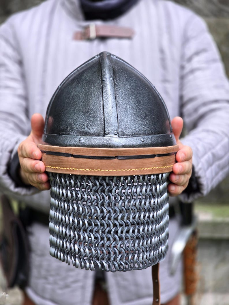 May include: A medieval-style helmet with a dark, metallic finish. The helmet features a rounded top and a chainmail lower section. A brown leather band encircles the helmet. The helmet is held by a person wearing a gray padded garment.