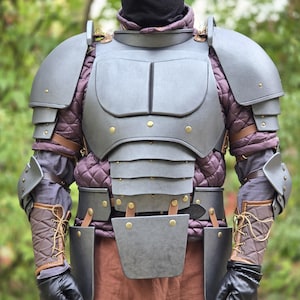 Puede incluir: Un conjunto de armadura de placas gris oscuro que incluye una coraza, hombreras y protectores de brazo, sobre un jubón acolchado morado. La armadura se asegura con correas de cuero marrón y remaches de color latón, junto con guantes negros.