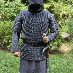 May include: A person wearing a medieval chainmail shirt, a black hood, and a gray tunic. A leather belt with a buckle is around the waist. The person is holding a sword with a brown leather-wrapped handle. The background is green foliage.