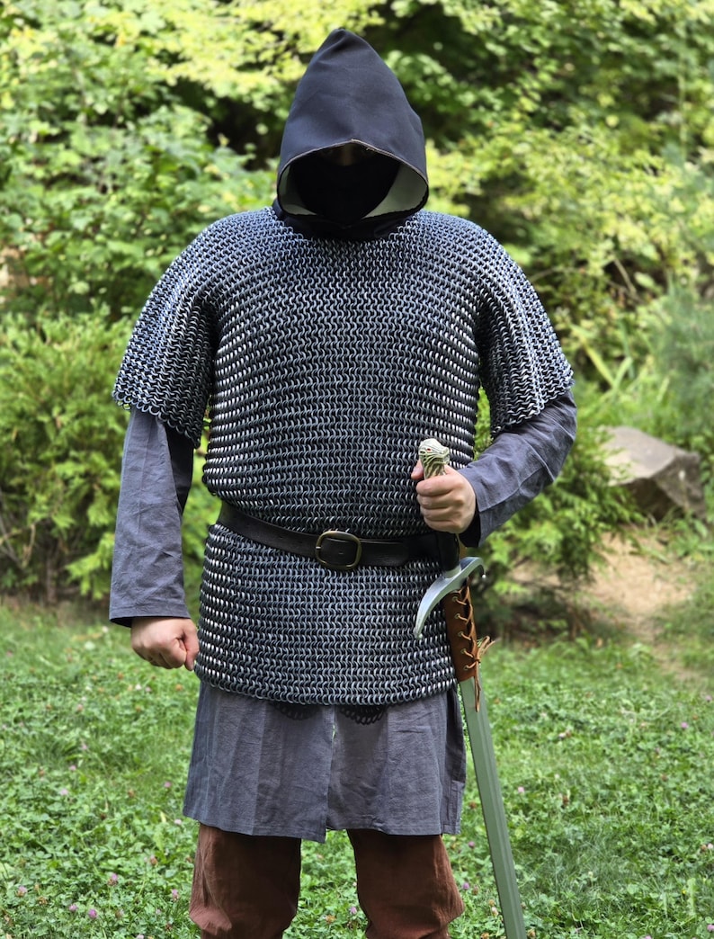 May include: A person wearing a medieval chainmail shirt, a black hood, and a gray tunic. A leather belt with a buckle is around the waist. The person is holding a sword with a brown leather-wrapped handle. The background is green foliage.