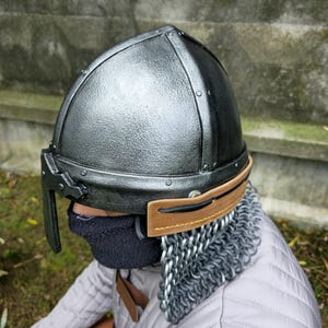 May include: A silver and black medieval helmet with riveted construction. The helmet features a brown leather visor and a chainmail neck guard. The person is wearing a quilted gray jacket.
