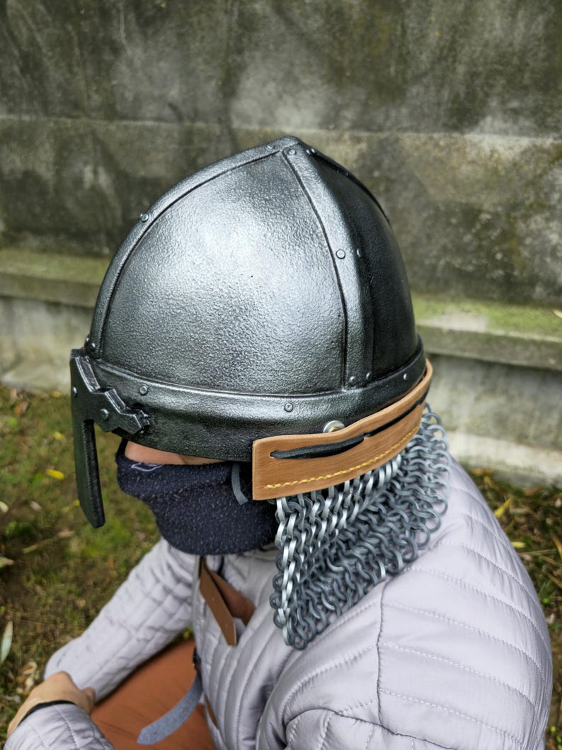 May include: A silver and black medieval helmet with riveted construction. The helmet features a brown leather visor and a chainmail neck guard. The person is wearing a quilted gray jacket.