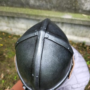 May include: A black medieval helmet with riveted metal bands. The helmet has a textured surface and a rounded top, suggesting a historical or fantasy theme. The design includes a cross-shaped reinforcement.