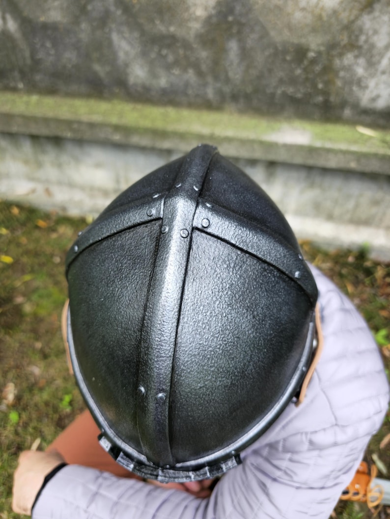 May include: A black medieval helmet with riveted metal bands. The helmet has a textured surface and a rounded top, suggesting a historical or fantasy theme. The design includes a cross-shaped reinforcement.
