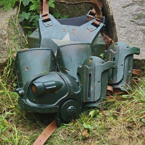 May include: A set of dark green armor pieces with brown leather straps. The armor includes a chest plate, shoulder guards, and helmet-like components. A white star is visible on the chest plate. The armor is resting on grass and near a large rock.