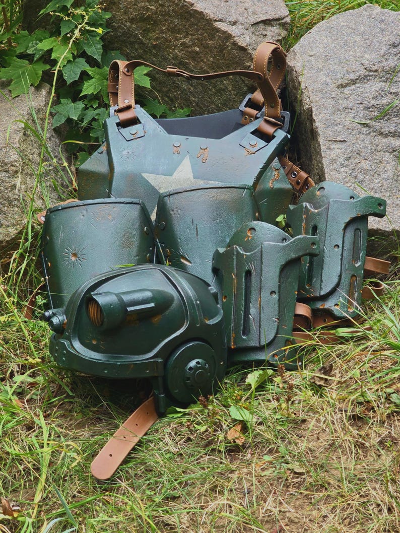 May include: A set of dark green armor pieces with brown leather straps. The armor includes a chest plate, shoulder guards, and helmet-like components. A white star is visible on the chest plate. The armor is resting on grass and near a large rock.