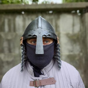 May include: A medieval-style helmet with a cross-shaped visor and chainmail. The helmet is black with a silver cross and brown leather accents. The person is wearing a gray quilted jacket with a brown leather strap.