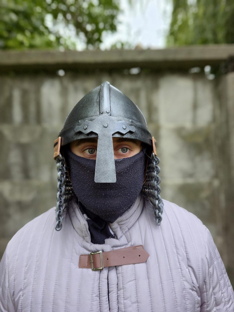 May include: A medieval-style helmet with a cross-shaped visor and chainmail. The helmet is black with a silver cross and brown leather accents. The person is wearing a gray quilted jacket with a brown leather strap.