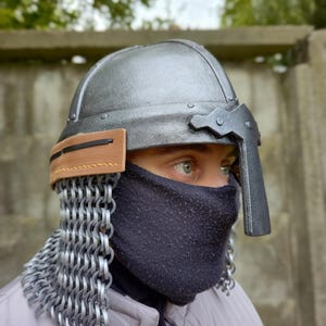 May include: A silver-colored medieval helmet with a visor and a leather strap. The helmet has a chainmail neck guard and a black face covering. The person is wearing a light gray quilted jacket with a brown leather strap.