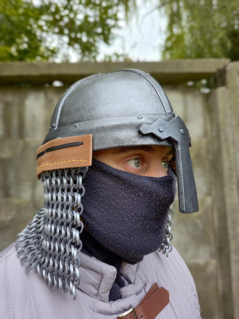 May include: A silver-colored medieval helmet with a visor and a leather strap. The helmet has a chainmail neck guard and a black face covering. The person is wearing a light gray quilted jacket with a brown leather strap.