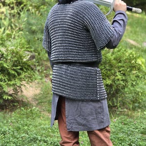 May include: A person in medieval attire, including a chainmail shirt, hooded cloak, and brown boots, holds a large silver sword over their shoulder. The outfit also includes brown pants and a gray tunic. The background is a lush green environment.