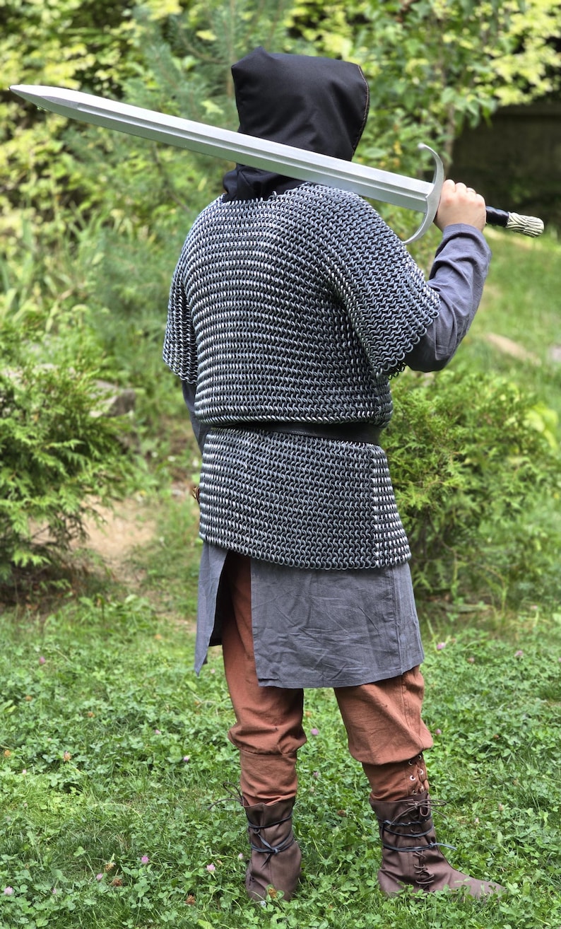 May include: A person in medieval attire, including a chainmail shirt, hooded cloak, and brown boots, holds a large silver sword over their shoulder. The outfit also includes brown pants and a gray tunic. The background is a lush green environment.