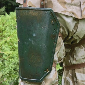 May include: A green, riveted metal arm guard with a distressed finish. The arm guard is attached to the leg with brown leather straps. The person is wearing camouflage pants and a matching jacket.