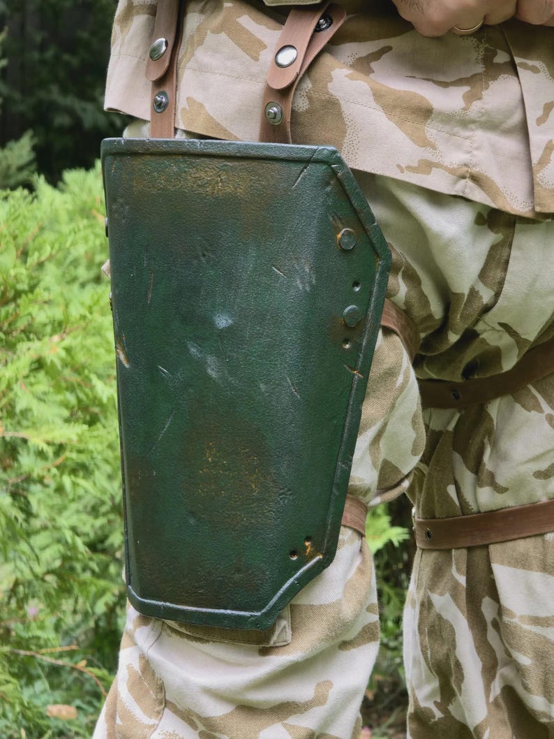 May include: A green, riveted metal arm guard with a distressed finish. The arm guard is attached to the leg with brown leather straps. The person is wearing camouflage pants and a matching jacket.
