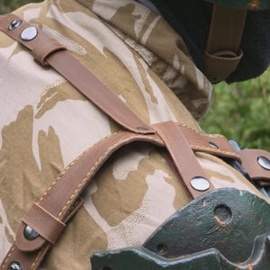 May include: Close-up of a brown leather harness with silver rivets, worn over a camouflage jacket. The jacket has a tan and brown pattern. A green piece of equipment is visible on the right side of the image.