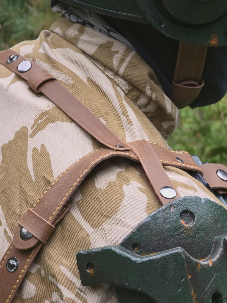 May include: Close-up of a brown leather harness with silver rivets, worn over a camouflage jacket. The jacket has a tan and brown pattern. A green piece of equipment is visible on the right side of the image.