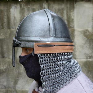 May include: A silver-colored medieval helmet with a chainmail neck guard. The helmet has a rounded top and a visor. A brown leather band is attached to the helmet. The person is wearing a black face covering and a light-colored quilted garment.