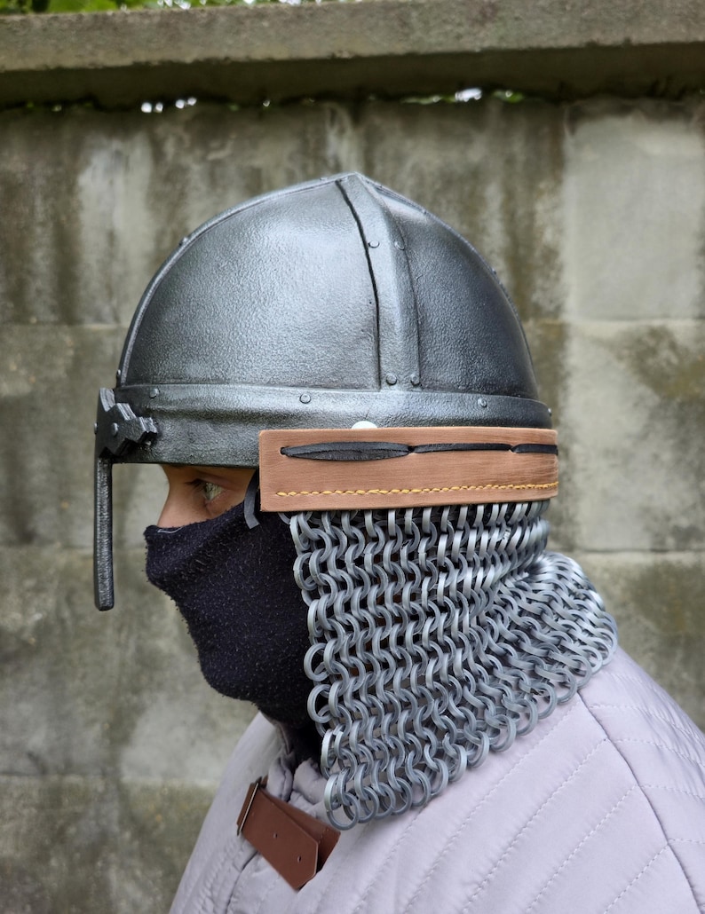 May include: A silver-colored medieval helmet with a chainmail neck guard. The helmet has a rounded top and a visor. A brown leather band is attached to the helmet. The person is wearing a black face covering and a light-colored quilted garment.