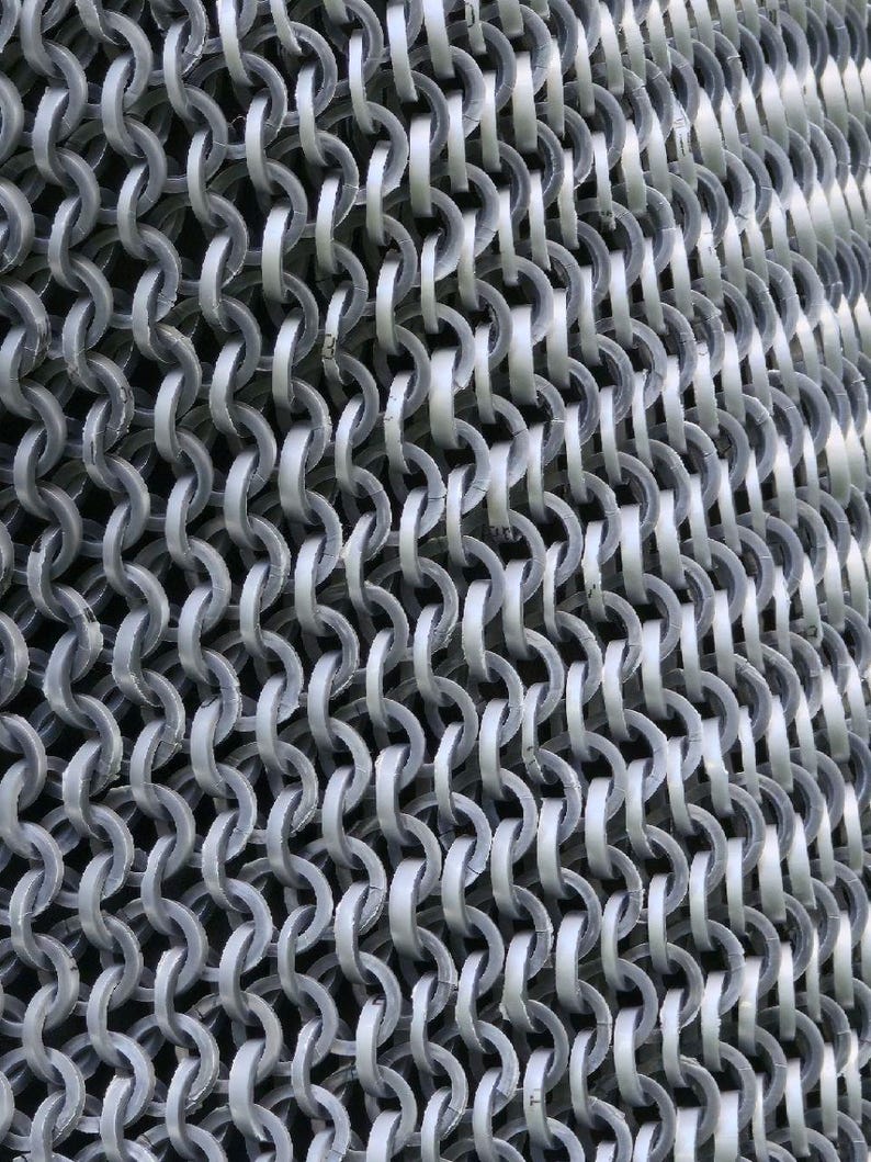 May include: Close-up of a chainmail texture. The image shows a dense pattern of interlocking, silvery-gray metal rings. The rings are arranged in rows and columns, creating a flexible, mesh-like structure. The lighting highlights the metallic sheen.