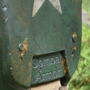 May include: Close-up of a green combat armor shield with a white star design. The shield has the words "Combat Armor" embossed on the lower section. The shield has a distressed, weathered appearance with visible scratches and rust-colored accents.