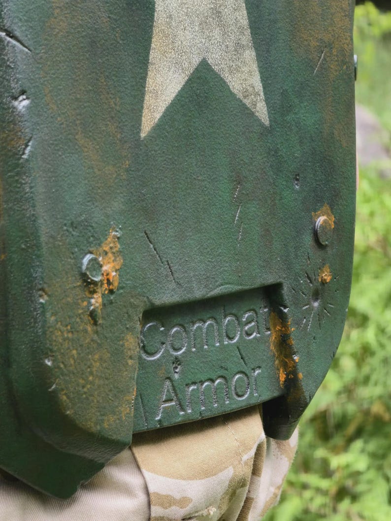 May include: Close-up of a green combat armor shield with a white star design. The shield has the words "Combat Armor" embossed on the lower section. The shield has a distressed, weathered appearance with visible scratches and rust-colored accents.