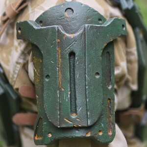 May include: A weathered, dark green, rectangular object with a curved top and multiple holes and slots. The object has a distressed, textured appearance with hints of orange. It is attached to a tan and brown camouflage fabric with leather straps.