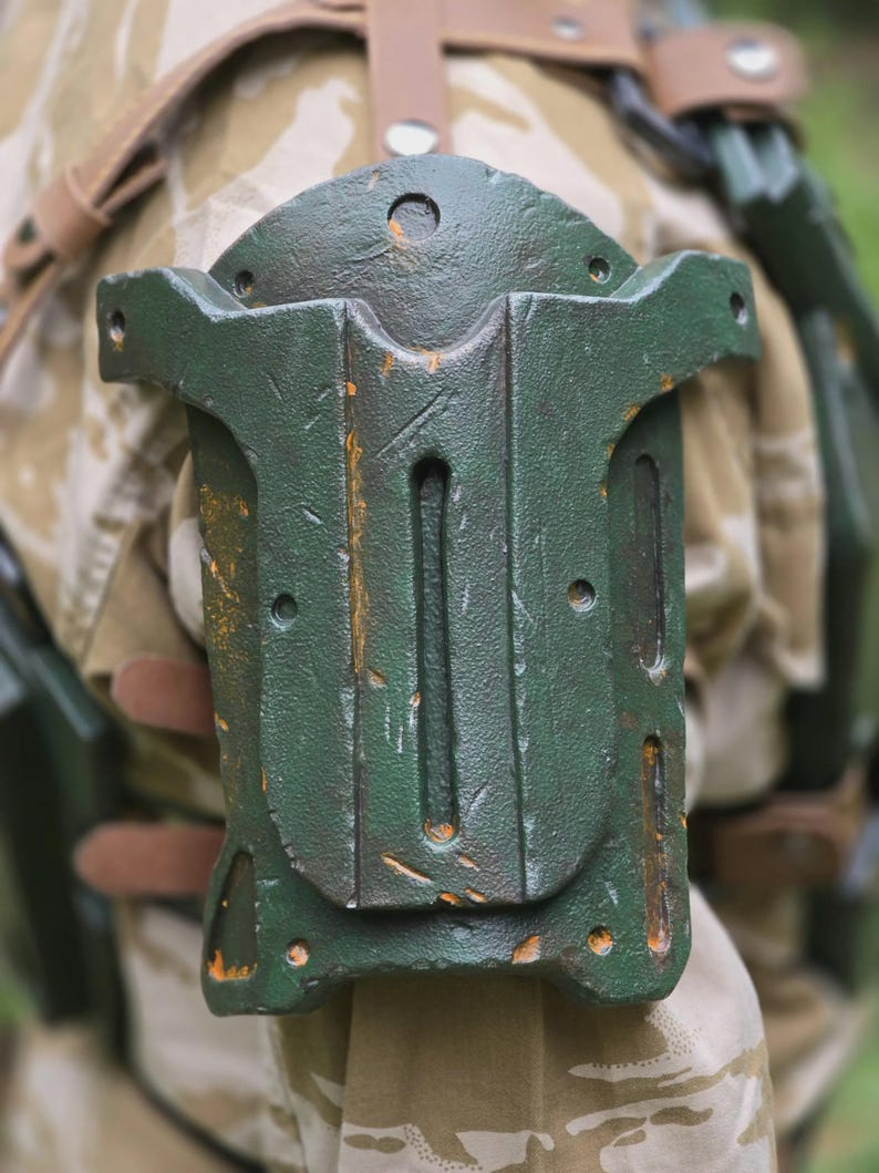 May include: A weathered, dark green, rectangular object with a curved top and multiple holes and slots. The object has a distressed, textured appearance with hints of orange. It is attached to a tan and brown camouflage fabric with leather straps.