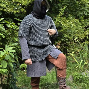 May include: A person wearing a medieval-style outfit, including a chainmail shirt, a black hood, and brown pants. The outfit also includes a gray tunic and brown leather boots. The person is standing in a natural outdoor setting.