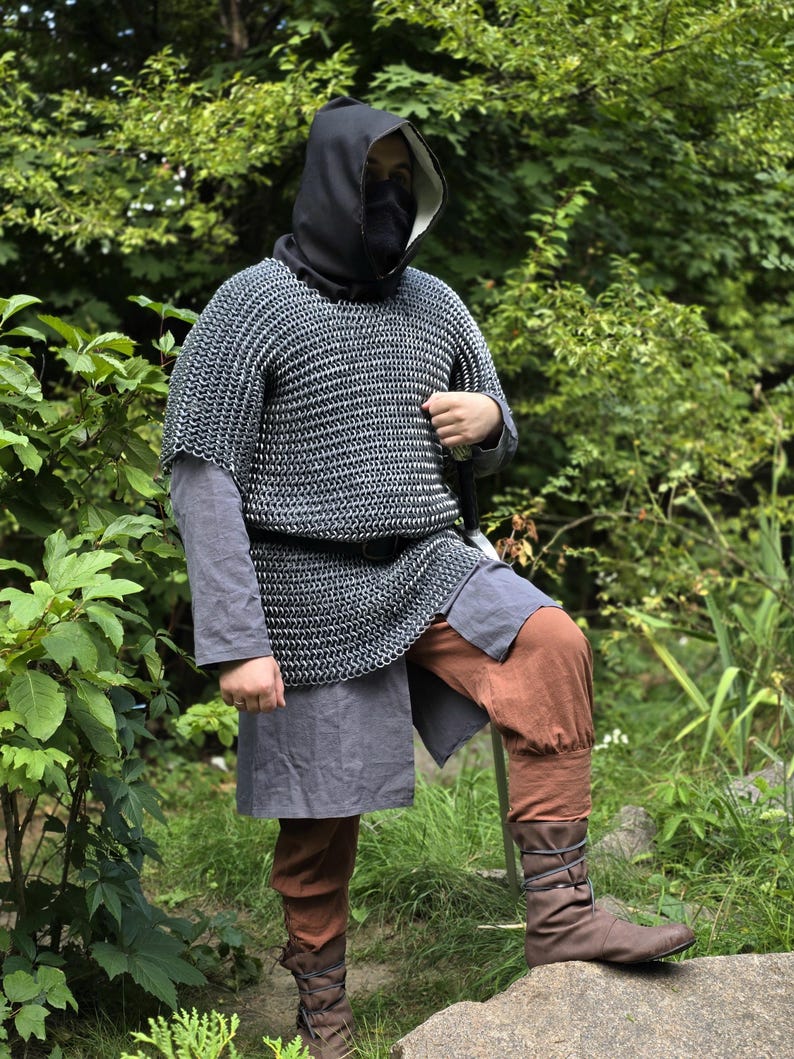 May include: A person wearing a medieval-style outfit, including a chainmail shirt, a black hood, and brown pants. The outfit also includes a gray tunic and brown leather boots. The person is standing in a natural outdoor setting.