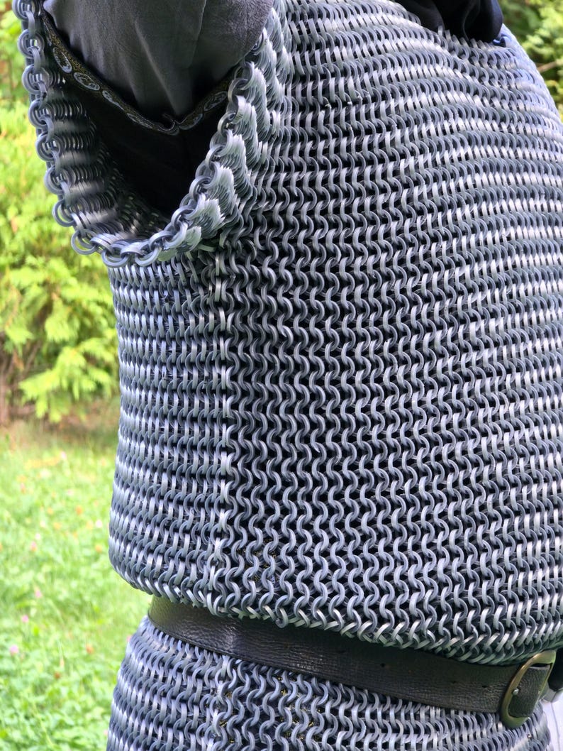 May include: Close-up of a chainmail shirt, a type of armor made of interlocking metal rings. The shirt is silver and gray, with a black leather belt. The background is blurred green foliage.