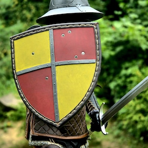 Op de afbeelding: Een middeleeuws schild met een gevierendeeld ontwerp van gele en rode vlakken, gescheiden door een grijs metalen kruis. Het schild heeft een donkerbruine, gestikte lederen rand. Een grijze metalen helm en een zwaard zijn ook zichtbaar.