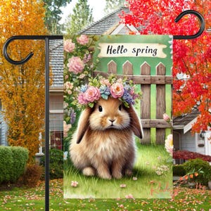 May include: A garden flag featuring a brown bunny wearing a flower crown. The bunny is seated in front of a wooden fence with a green background. The text "Hello Spring" is displayed on a sign above the fence.