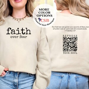 May include: A beige sweatshirt with the text "faith over fear" printed on the front. The back of the sweatshirt has a QR code with the text "REFRESH YOUR SOUL" and a Bible verse from 2 Timothy 1:7.