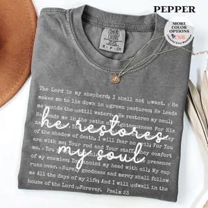 Puede incluir: Una camiseta de cuello redondo gris carb&oacute;n que presenta el texto completo del Salmo 23 impreso en fuente blanca peque&ntilde;a, con la frase he restores my soul escrita en una fuente cursiva blanca m&aacute;s grande en el centro. Etiqueta Comfort Colors.