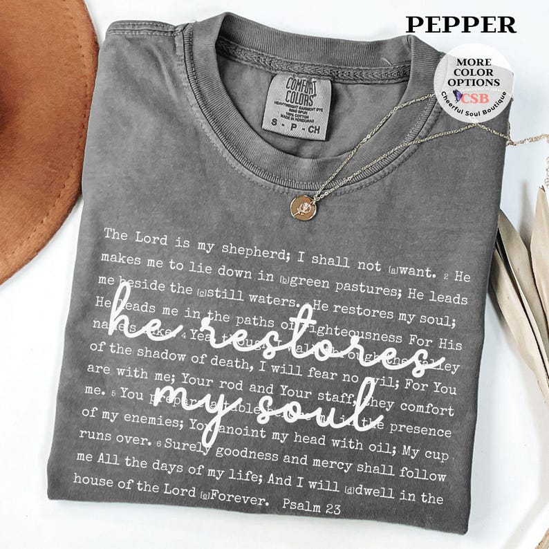 Puede incluir: Una camiseta de cuello redondo gris carb&oacute;n que presenta el texto completo del Salmo 23 impreso en fuente blanca peque&ntilde;a, con la frase he restores my soul escrita en una fuente cursiva blanca m&aacute;s grande en el centro. Etiqueta Comfort Colors.